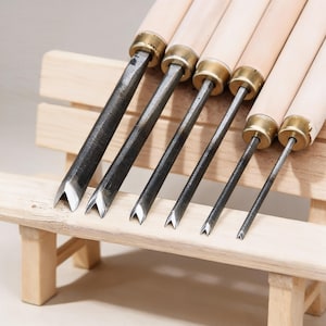 Może przedstawiać: Zestaw sześciu dłut do rzeźbienia w drewnie z drewnianymi uchwytami i metalowymi ostrzami. Ostrza są kątowe i mają krawędź tnącą w kształcie litery V. Dłuta są ułożone na małej drewnianej ławce.