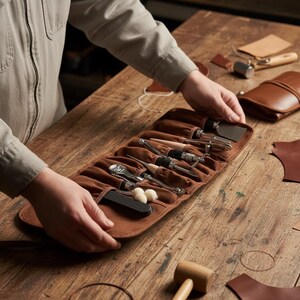 May include: A brown leather tool roll with various metal and wooden tools. The roll is open, displaying the tools, and is held by a person. Other leather tools and materials are on the wooden surface.