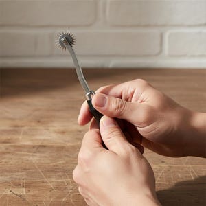 May include: A silver and black dressmaker's tracing wheel with a spiked wheel at the top. The handle is black and the wheel is held in place by a curved metal arm. The tool is designed for transferring pattern markings onto fabric.