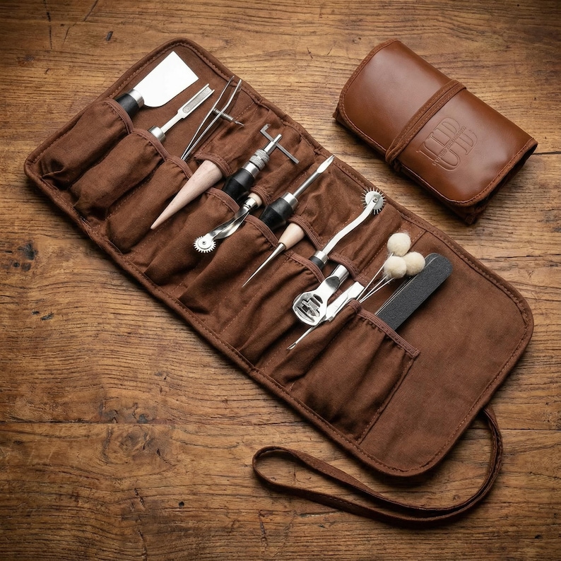 May include: A brown leather tool roll with various metal tools, including a spatula, tweezers, and awls. The roll is open, displaying the tools in individual pockets. A matching leather case with embossed initials is next to the roll. The tools are on a wooden surface.