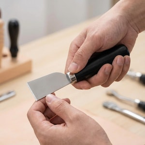 May include: A leatherworking tool with a black handle and a silver blade. The tool is held in two hands, with other leatherworking tools visible in the background. The blade is rectangular and appears sharp.