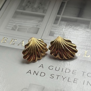 May include: A pair of gold-colored seashell-shaped stud earrings. The earrings have a detailed, ridged design and a polished finish. The earrings are displayed on a book with the text "A GUIDE TO AND STYLE IN".