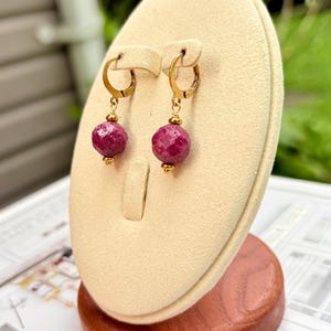 May include: A pair of gold-tone hoop earrings featuring faceted, round, deep pink gemstone beads. Each earring is accented with small gold-tone metal spacers above and below the bead, suspended from a hinged gold-tone hoop closure mechanism.