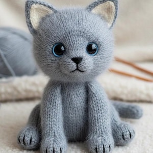 Peut inclure: Un chat gris tricoté à la main avec de grands yeux bleus. Le jouet en peluche a un nez et des pattes noirs et est assis droit. Une pelote de laine grise et des aiguilles à tricoter sont en arrière-plan.
