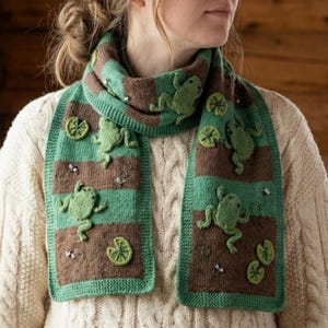 May include: A green and brown knitted scarf with frog and lily pad appliqués. The scarf features alternating stripes of green and brown, with detailed frog and lily pad designs. The scarf is worn over a cream-colored sweater.