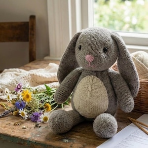 May include: A handmade gray knitted bunny with long ears, a pink nose, and a cream-colored belly. The bunny sits on a wooden table next to a bouquet of wildflowers and a basket of yarn.