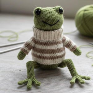 Könnte beinhalten: Eine handgefertigte grüne Strickfroschpuppe mit einem cremefarbenen und braun gestreiften Pullover. Der Frosch hat schwarze Augen und sitzt mit ausgestreckten Beinen. Stricknadeln und ein Knäuel grüne Wolle befinden sich im Hintergrund.
