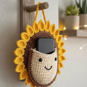 May include: A handmade crochet sunflower wall pocket with a smiling face design. The pocket is tan with a brown border and yellow petals. A black smartphone is inside the pocket. The pocket is hanging on a wooden wall mount.