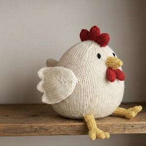 Può includere: Un giocattolo a forma di pollo bianco lavorato a maglia con cresta e bargigli rossi, becco e zampe gialle e occhi neri. Il pollo è seduto su una mensola di legno. Le ali sono di una tonalità di bianco più chiara.