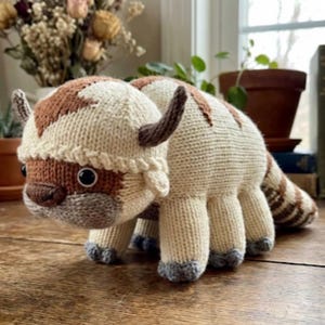 May include: A handmade, knitted plush toy in the shape of a bison. The toy features a cream-colored body with brown and tan accents on the head, tail, and hooves. It has small, dark eyes and curved horns.