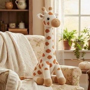 Puede incluir: Un juguete de jirafa tejido con cuerpo blanco y manchas marrones está sentado en un sillón floral. La jirafa tiene un cuello largo, orejas pequeñas y un hocico marrón. Una manta color crema está sobre la silla. Una pequeña mesa tiene lana y agujas de tejer.