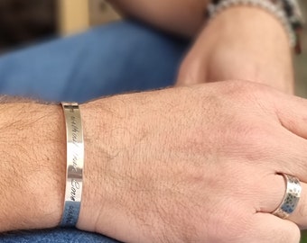 May include: A silver-toned metal cuff bracelet featuring engraved cursive text along its outer surface. The bracelet has a smooth, polished finish and a slim, open-ended design that rests securely around the wrist, showcasing clear lettering.