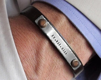 May include: A dark brown leather bracelet with a silver metal plate engraved with the roman numerals IV.VII.XIII. The bracelet has copper-colored rivets and is worn on a wrist, with a light-colored blazer and blue shirt cuff in the background.