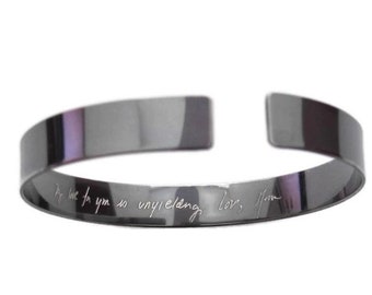 Puede incluir: Una pulsera tipo brazalete de color gris plomo pulido con un diseño abierto. La superficie interior presenta un mensaje grabado en letra cursiva que dice: My love for you is unyielding, love, Mom. El metal tiene un acabado liso y brillante.