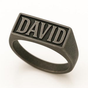 May include: A dark gray signet ring with the name "DAVID" in bold, raised letters. The ring has a rectangular face and a smooth, rounded band. The ring is made of a matte metal.