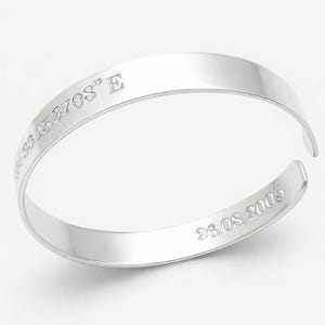Puede incluir: Un brazalete rígido de tono plateado pulido con diseño abierto. La superficie exterior está grabada con las coordenadas 48°36'15.3708" E, mientras que la superficie interior muestra la fecha grabada 26.08.2005 en una fuente sans-serif.
