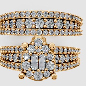 Könnte beinhalten: Zweiteiliges, goldfarbenes Ring-Set. Die Ringe sind mit zahlreichen kleinen, runden, klaren Edelsteinen besetzt. Der Hauptring hat eine tropfenförmige Anordnung größerer, klarer Edelsteine mit zwei rechteckigen Steinen in der Mitte.