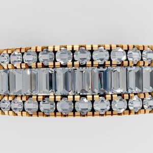 Puede incluir: Un anillo dorado con tres filas de piedras preciosas transparentes. La fila central tiene piedras rectangulares, flanqueadas por filas de piedras redondas. El anillo es una joya.