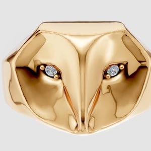 May include: A gold-colored ring with a geometric owl face design. The ring features two small, sparkling gemstones set as eyes. The ring has a polished, reflective surface, and the owl's face is the primary design element.
