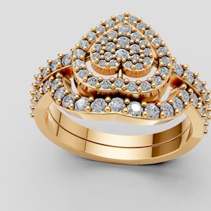 Puede incluir: Un juego de anillos dorados con un diseño en forma de corazón. El anillo superior está adornado con numerosas piedras preciosas pequeñas y transparentes, y la banda también está adornada con piedras preciosas. El conjunto incluye una banda a juego.