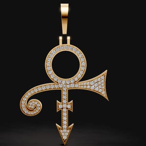 May include: A gold-colored pendant in the shape of a stylized symbol, encrusted with numerous small, sparkling gemstones. The pendant has a loop at the top for a chain and a downward-pointing arrow at the bottom. The background is a dark, solid color.