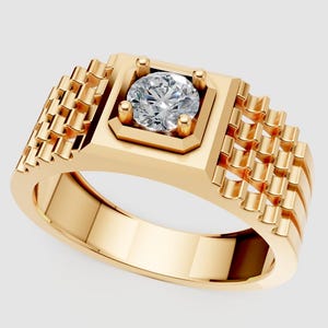 Puede incluir: Un anillo dorado con un diseño cuadrado y texturizado en los laterales. El anillo presenta una gema central, redonda y transparente, engastada en un bisel cuadrado. La banda es lisa y pulida.