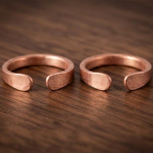 Könnte beinhalten: Zwei offene Ringe aus Kupfer mit einer ausgeprägten gehämmerten Textur auf der Außenfläche. Jeder Ring hat ein abgerundetes, glattes Ende an der Öffnung und zeigt den metallischen, rotbraunen Farbton des Kupfers.