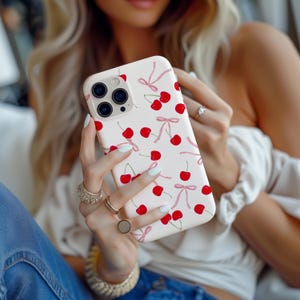 May include: A light pink phone case featuring a pattern of red cherries and pink bows. The case is held by a person wearing rings and a bracelet. The phone case has a camera bump with three lenses.