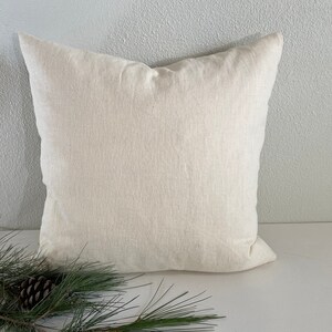 May include: A plain white linen pillow cover with a subtle texture. The pillow is sitting on a white surface with a pine branch in the foreground.