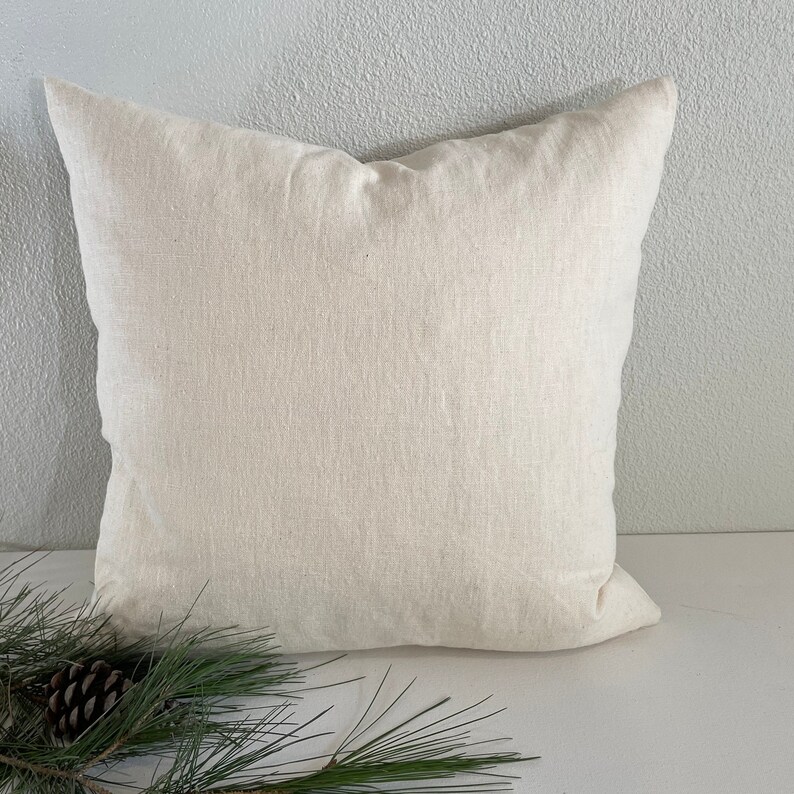 May include: A plain white linen pillow cover with a subtle texture. The pillow is sitting on a white surface with a pine branch in the foreground.