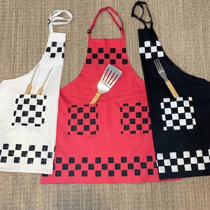 May include: Three aprons in white, red, and black, each featuring a checkered pattern on the pocket and hem. The white apron has two wooden-handled tools in the pocket, the red apron has a spatula, and the black apron has a fork.