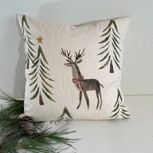 May include: A white cushion with a beige background features a stylised reindeer with a red bow and green pine trees. The cushion has a gold star in the top left corner.