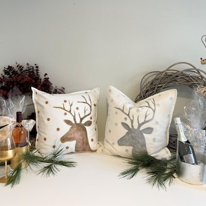 May include: Two white throw pillows with gold and silver reindeer silhouettes, gold and silver polka dots, and red accents. The pillows are on a white table with a bottle of wine, two gold wine glasses, and a silver ice bucket with a bottle of champagne.