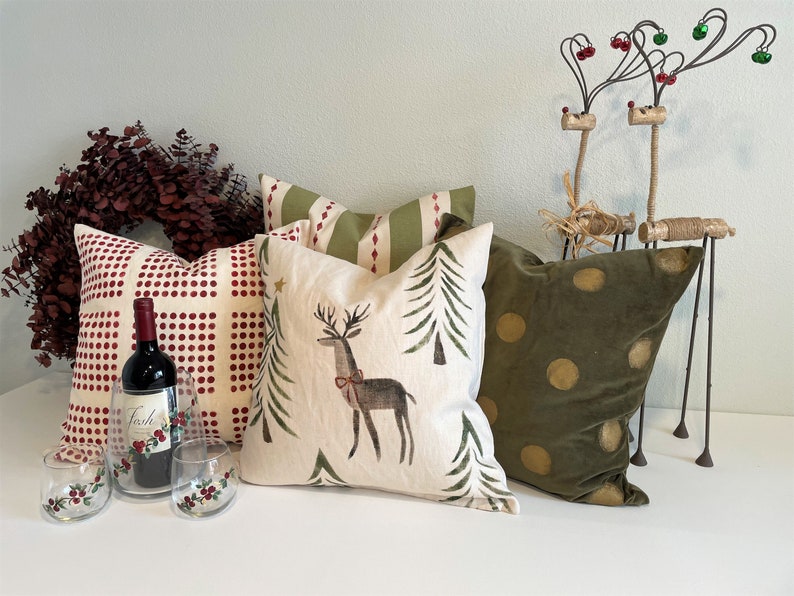 May include: A collection of decorative throw pillows with a Christmas theme. The pillows feature a red and white checked pattern, a green and white tree pattern, a cream pillow with a reindeer and trees, and a green velvet pillow with gold polka dots.