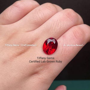 Puede incluir: Un rubí cultivado en laboratorio certificado, de forma ovalada y color rojo intenso. La gema está facetada para reflejar la luz. La imagen incluye el texto "Tiffany Gems | Craft jewellers" y "Certified Lab Grown Ruby".