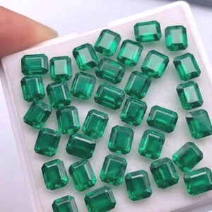 May include: A collection of emerald-cut emerald gemstones, each with a vibrant green hue. The gemstones are displayed in a white tray, showcasing their rectangular shape and facets. The image highlights the clarity and color of the precious stones.