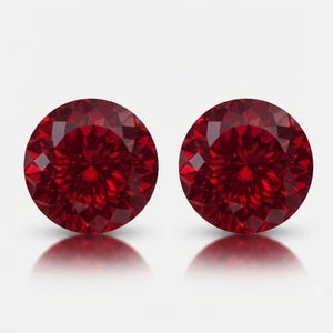 May include: Two round-cut, deep red gemstones with a faceted surface. Each stone displays a brilliant, symmetrical pattern of triangular and kite-shaped facets that reflect light, creating a vibrant, saturated crimson appearance against a plain background.