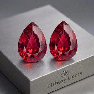 May include: Two faceted, teardrop-shaped, red gemstones. The gems are displayed on a brushed metal surface. The text "Tiffany Gems" is engraved on the metal surface.