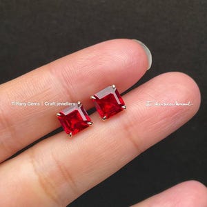 Puede incluir: Un par de pendientes de botón cuadrados con piedras preciosas rojas vibrantes y engastes dorados. Los pendientes se muestran sobre un fondo claro. El texto "Tiffany Gems | Craft jewellers" es visible.