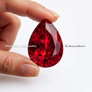 May include: A large, faceted, pear-shaped, deep red gemstone. The gem is held between fingers against a white background. The text "Tiffany Gems | Craft jewellers" and "The decision moment" are visible.