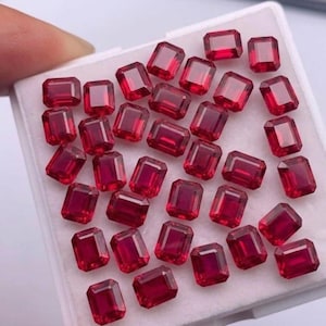 May include: A collection of rectangular-cut, deep red gemstones, possibly rubies, arranged in a white tray. The gems are faceted, reflecting light, and appear to be of high quality. The image is well-lit, showcasing the color and clarity of the stones.