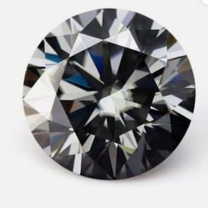 Puede incluir: Una gema facetada de corte redondo con un tono gris oscuro. La superficie de la gema refleja la luz, creando un juego de facetas brillante. La gema se encuentra sobre un fondo blanco, resaltando su claridad.