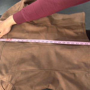 May include: A brown jacket featuring vertical seam detailing and a smooth texture. A pink measuring tape is placed horizontally across the back, indicating a width measurement of approximately 63.5 cm from edge to edge.