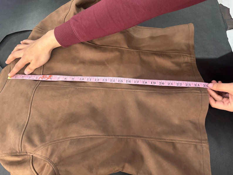 May include: A brown jacket featuring vertical seam detailing and a smooth texture. A pink measuring tape is placed horizontally across the back, indicating a width measurement of approximately 63.5 cm from edge to edge.