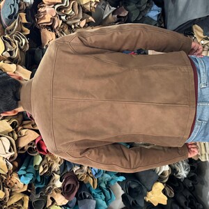May include: A brown suede jacket with a structured back panel, long sleeves, and a collar, worn over a dark red shirt and blue denim jeans. The jacket features visible vertical seam detailing along the back and a smooth, matte finish.