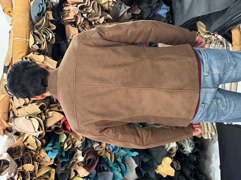 May include: A brown suede jacket with a structured back panel, long sleeves, and a collar, worn over a dark red shirt and blue denim jeans. The jacket features visible vertical seam detailing along the back and a smooth, matte finish.