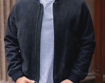 May include: A navy blue bomber jacket with a zip closure and ribbed cuffs. The jacket is worn over a grey t-shirt. The person is wearing black trousers and has their hands in their pockets.