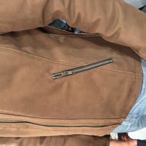 May include: A brown jacket with a smooth, matte finish featuring a long-sleeve design, a full-length front zip, and a diagonal zipped pocket on the side. The garment is constructed with visible seam detailing and dark metal hardware.