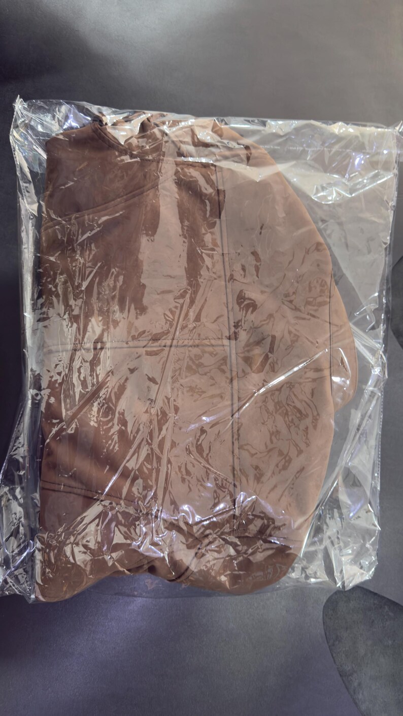 May include: A solid brown garment with visible stitched seams and a rounded neckline, encased in a clear, crinkled plastic protective bag. The fabric appears smooth and matte, with the packaging creating reflective highlights across the surface.
