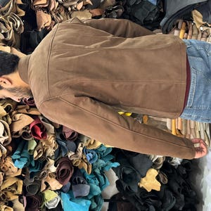 May include: A brown suede jacket with a structured collar and long sleeves, worn over a dark red shirt and blue denim jeans. The jacket features visible seam detailing along the back and shoulders, providing a clean and fitted appearance.